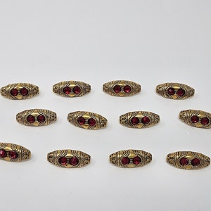 May include: Twelve gold-toned oval-shaped beads with red rhinestone accents. The beads have a decorative design.