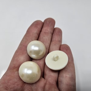 Op de afbeelding: Drie grote, ronde, crèmekleurige knopen met een parelmoerglans. Eén knoop toont de achterkant met een kleine schacht. Knoppen voor naai- of knutselprojecten.