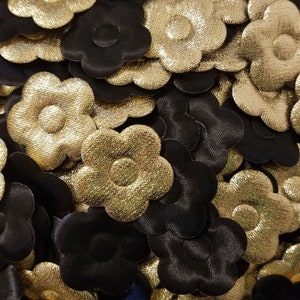 Può includere: Primo piano di un mucchio di decorazioni floreali in tessuto nero e oro. I fiori sono tutti della stessa dimensione e forma, e hanno un centro leggermente rialzato.