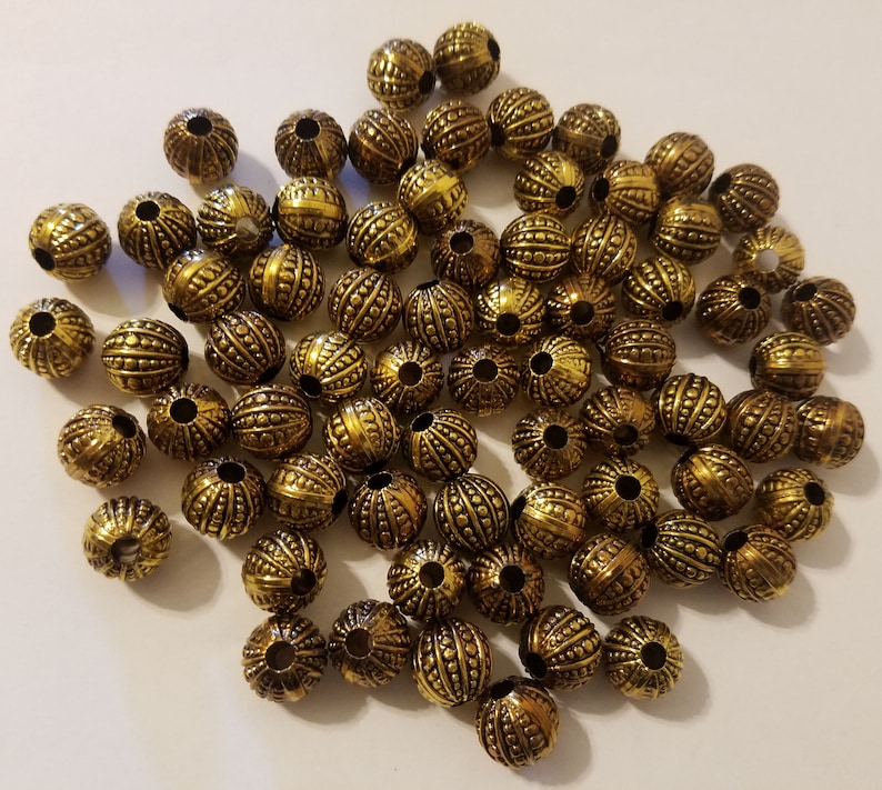 May include: A large group of gold-colored metal beads with a textured surface. The beads are round and have a hole in the center for stringing.