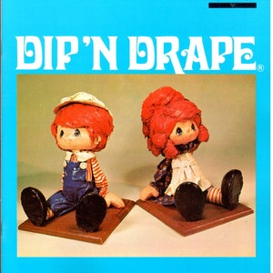 Puede incluir: Una portada de libro con el título "Dip 'N Drape" en letras blancas sobre un fondo azul. La portada presenta dos muñecas, un niño y una niña, sentados sobre bases de madera. Las muñecas están hechas de tela y tienen el pelo rojo y caras pintadas.