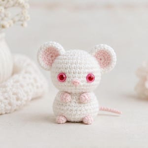 Puede incluir: Un peque&ntilde;o rat&oacute;n de ganchillo blanco con orejas grandes y redondas con centros de color rosa claro. El rat&oacute;n tiene ojos rosas brillantes, una nariz peque&ntilde;a de color rosa, patas rosas peque&ntilde;as y una cola rosa a juego, con textura de punto.