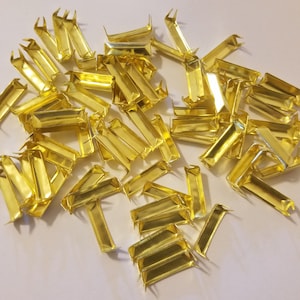 Puede incluir: Un montón de extremos de correa de metal de color dorado. Los extremos de la correa son rectangulares con dos pequeñas puntas en un extremo.