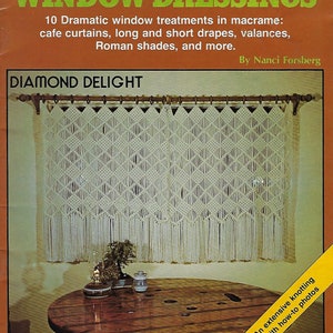 May include: A book cover with a white macrame window treatment, titled "Macrame Window Dressings: Diamond Delight". The book cover features a photo of a white macrame window treatment hanging in a room with a wooden table and a small plant. The book cover also includes the text "10 Dramatic window treatments in macrame: cafe curtains, long and short drapes, valances, Roman shades, and more." and "By Nanci Forsberg".
