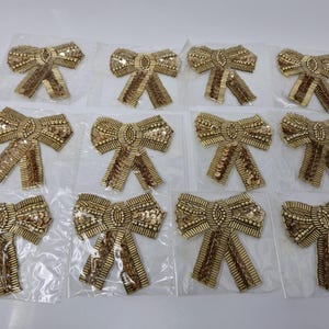 Può includere: Dodici applicazioni in tessuto a forma di fiocco di colore oro con paillettes e perline. Le applicazioni sono confezionate singolarmente su fogli di plastica trasparente.