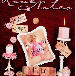 May include: A book titled "Love Notes" with a pink and white angel design on the cover. The book features a variety of decorative items, including a pillow, candles, and small decorative items with the words "laughter", "garden", "heart to heart", "old flames", and "love knots".