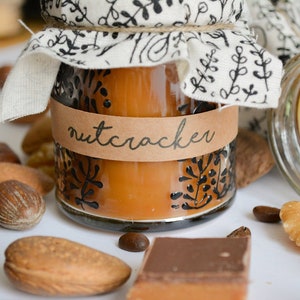 Può includere: Un barattolo di vetro con un coperchio marrone e un'etichetta marrone su cui è scritto "nutcracker". Il barattolo è pieno di un liquido marrone e ha un rivestimento in tessuto bianco sul coperchio. Il barattolo è circondato da noci, chicchi di caffè e una barretta di cioccolato.