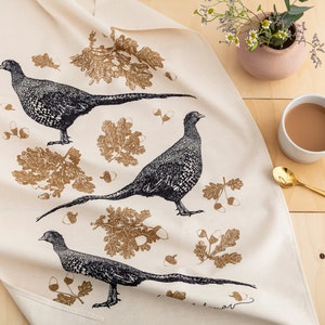 May include: A white tea towel with a black and white illustration of three pheasants and gold leaf designs. The tea towel is on a wooden surface with a cup of coffee and a gold spoon.