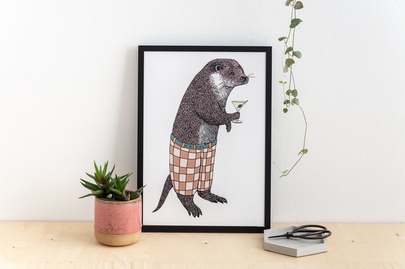 May include: A black framed print of a cartoon otter wearing plaid shorts and holding a martini glass. The otter is standing on a white background.