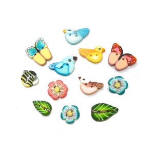 May include: A collection of decorative buttons in various shapes, including birds, butterflies, flowers, a bee, and leaves. The buttons are brightly colored with details like wings, petals, and stripes. The buttons are arranged on a white background.