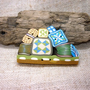 May include: A colourful brooch featuring miniature spools of thread and quilt block designs. The brooch has a brown base with green and white accents, and includes blue, green, and tan details. The brooch is approximately 5 cm wide.