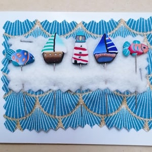 Op de afbeelding: Handgemaakte nautische wenskaart met een witte achtergrond en een blauw en goudkleurige patroonrand. De kaart toont miniatuur speldjes in de vorm van zeilboten, vuurtorens en vissen. De speldjes zijn in verschillende kleuren, waaronder blauw, rood en groen.