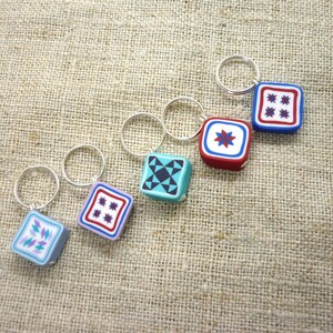 Op de afbeelding: Een verzameling van vijf vierkante sleutelhangers met kleurrijke geometrische patronen. Elke sleutelhanger heeft een zilveren ring en een kleine, kubusvormige bedel met een uniek ontwerp in tinten blauw, rood en teal. De sleutelhangers worden getoond op een getextureerde, bruinachtige stof.