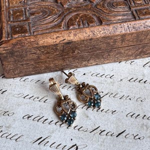 May include: A pair of drop earrings featuring a gold-toned metal filigree design in a heart-like shape, accented with three small, round, dark blue stones at the base of each earring, suspended from a simple metal hook attachment.