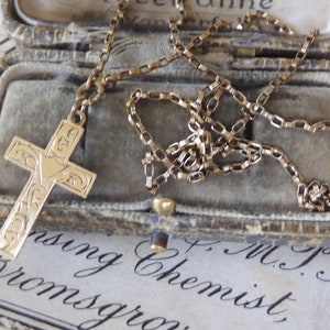 May include: A gold-coloured cross pendant necklace with an ornate design, inside a vintage grey velvet jewellery box. The chain is delicate and the cross features engraved details. The background includes faded text.