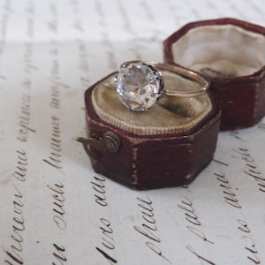 Può includere: Un anello di fidanzamento vintage con una grande gemma trasparente incastonata in una fascia d'oro, esposto in un portagioie bordeaux antico aperto con interno in velluto. Il portagioie è accanto a un altro simile.