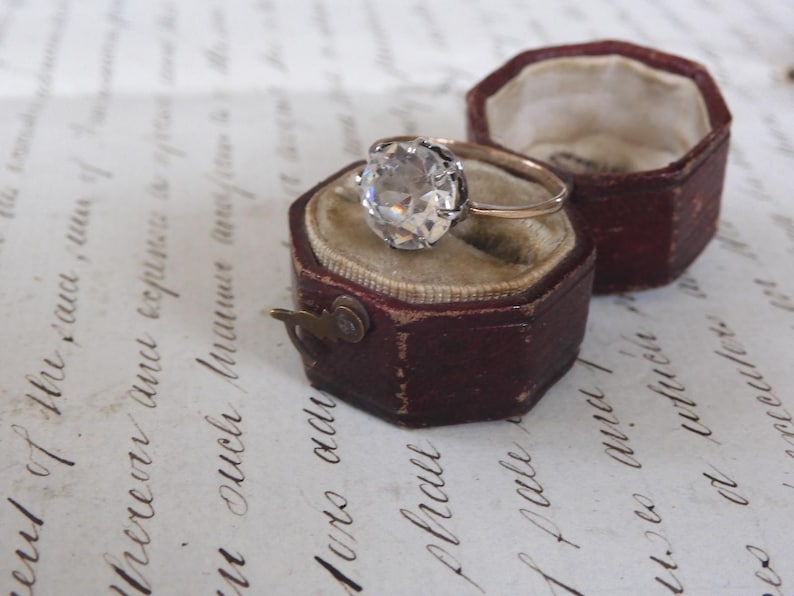 May include: A vintage engagement ring with a large, clear gemstone set in a gold band, displayed in an open, antique burgundy ring box with a velvet interior. The ring box is next to another similar box.
