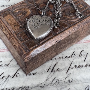 May include: A silver-toned heart-shaped locket pendant suspended from a matching delicate link chain. The locket features an engraved floral pattern on the upper portion of its surface, while the lower section remains smooth and polished.