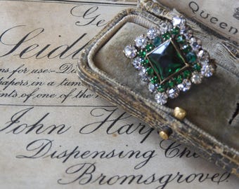 Könnte beinhalten: Eine vintage Brosche aus smaragdgrünem und klarem Strass in einer samtenen Box. Die Brosche hat die Form eines Quadrats mit einem großen smaragdgrünen Stein in der Mitte.
