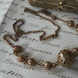May include: A vintage gold-toned necklace featuring a series of openwork spheres and a larger central orb. The necklace is displayed on a handwritten document, showcasing its intricate design and antique aesthetic.