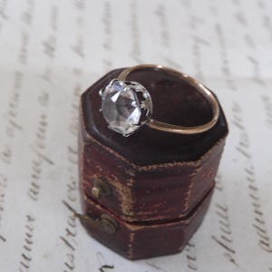 May include: A gold ring with a large, clear gemstone set in a silver-colored bezel. The ring rests on a small, antique, burgundy-colored jewelry box with a brass clasp. The background features handwritten script.