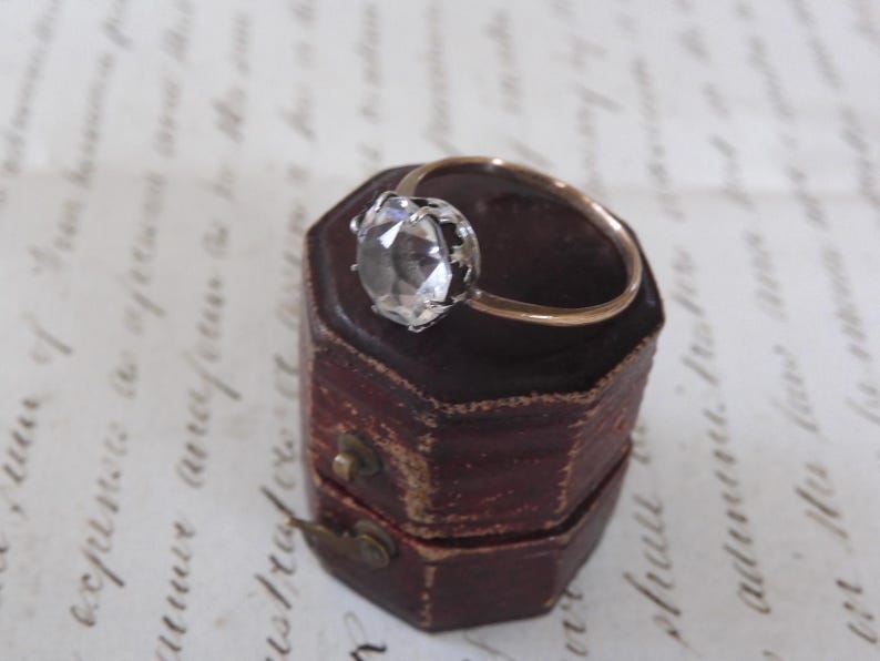 May include: A gold ring with a large, clear gemstone set in a silver-colored bezel. The ring rests on a small, antique, burgundy-colored jewelry box with a brass clasp. The background features handwritten script.