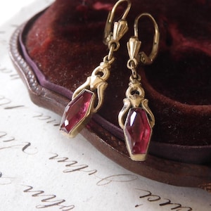 May include: A pair of antique gold-toned earrings with faceted, rectangular, deep red gemstones. The earrings are displayed against a dark burgundy velvet background, suggesting a vintage jewelry theme.