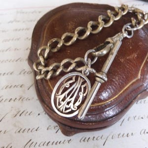 May include: An antique silver-toned watch chain with an oval pendant featuring an ornate monogram. The chain is connected to a T-bar and rests on a brown leather box. The background is a vintage paper.
