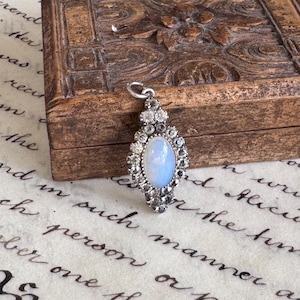 May include: A silver-toned pendant featuring a central oval-shaped iridescent cabochon stone surrounded by a border of small, clear faceted stones. The pendant has a small metal loop at the top for attachment and a pointed, elongated shape.