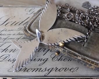 May include: A silver pendant necklace featuring two stylized bird wings. The wings are joined at the top and have a delicate, detailed design. The necklace is on a silver chain and is displayed in a velvet-lined box.