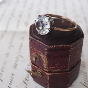 May include: A vintage-style ring with a large, faceted clear gemstone set in a dark metal bezel. The ring's band is gold-toned, and it rests on a small, antique burgundy jewelry box with a brass clasp. The background features handwritten script.