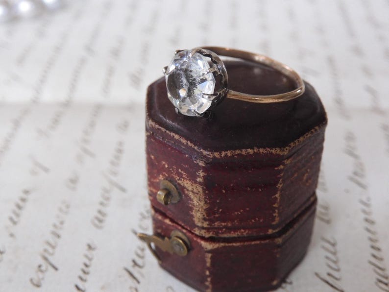May include: A vintage-style ring with a large, faceted clear gemstone set in a dark metal bezel. The ring's band is gold-toned, and it rests on a small, antique burgundy jewelry box with a brass clasp. The background features handwritten script.