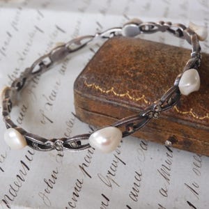 May include: A silver-toned bracelet with alternating links and large, irregular white pearls. The bracelet is resting on a vintage leather jewelry box with gold trim, and a handwritten paper.