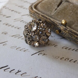 Op de afbeelding: Een vintage ring met een cluster van heldere, facetgeslepen stenen gezet in een aangetast zilverkleurig metaal. De ring is rond en ligt op verouderd papier met handschrift, naast een met fluweel gevoerde sieradendoos.