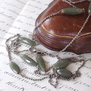 May include: A vintage necklace featuring oval-shaped, olive-green beads strung on a silver-tone chain. The necklace is displayed on a handwritten document and near a brown leather jewelry box. The necklace has a classic, elegant design.