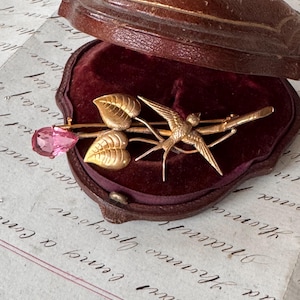 Op de afbeelding: Goudkleurige broche met een vogel op een tak, bladeren en een roze edelsteen. De broche wordt gepresenteerd in een vintage bruin leren sieradendoosje met een bordeauxrood fluwelen interieur.