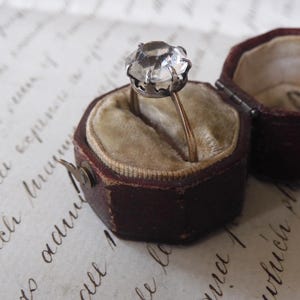 May include: A vintage ring with a large, faceted clear stone set in a silver-colored metal. The ring is displayed in an open, antique, burgundy-colored ring box with a beige velvet interior. The box is on a paper with handwritten script.