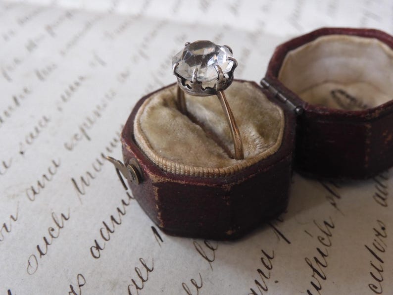 May include: A vintage ring with a large, faceted clear stone set in a silver-colored metal. The ring is displayed in an open, antique, burgundy-colored ring box with a beige velvet interior. The box is on a paper with handwritten script.