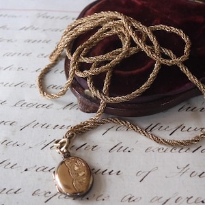 May include: A gold-toned necklace with a braided chain and a round pendant. The pendant has an engraved design and is attached to the chain with a small clasp. The necklace rests on a vintage paper with handwritten text.
