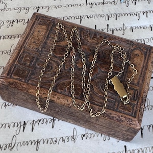 May include: A gold-toned metal chain necklace featuring a series of small, interconnected circular links and a single, smooth, elongated rectangular pendant with scalloped edges, resting on a dark brown, rectangular wooden box with carved details.