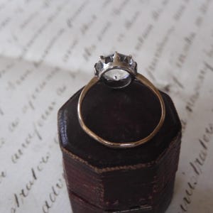 May include: A vintage-style ring with a clear, faceted gemstone set in a silver-colored crown. The ring has a thin, gold-colored band and rests on a dark brown, hexagonal jewelry box. The background features faded script.