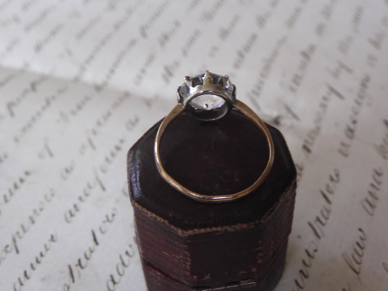 May include: A vintage-style ring with a clear, faceted gemstone set in a silver-colored crown. The ring has a thin, gold-colored band and rests on a dark brown, hexagonal jewelry box. The background features faded script.