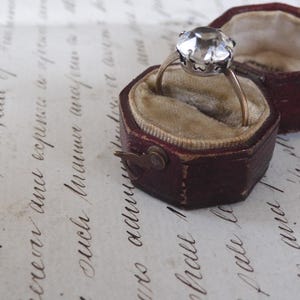 May include: A vintage ring with a large, faceted clear gemstone set in a silver-toned metal. The ring rests in an open, burgundy leather ring box with a velvet interior. The box is next to its matching lid.