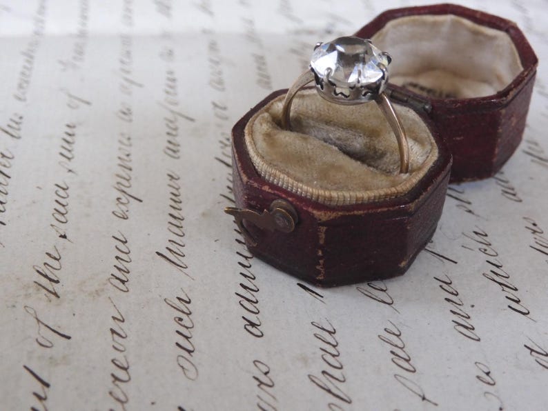 May include: A vintage ring with a large, faceted clear gemstone set in a silver-toned metal. The ring rests in an open, burgundy leather ring box with a velvet interior. The box is next to its matching lid.