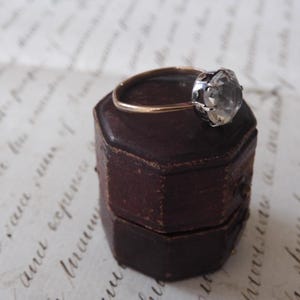 May include: A vintage ring with a large, clear gemstone set in a silver and gold band, resting on a small, antique, brown leather ring box. The ring box is hexagonal and has a worn, aged appearance.
