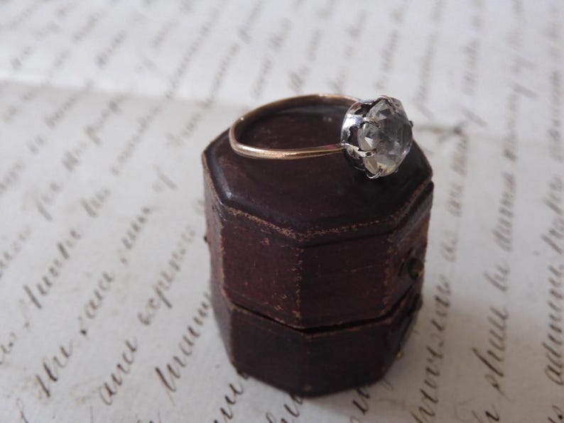 May include: A vintage ring with a large, clear gemstone set in a silver and gold band, resting on a small, antique, brown leather ring box. The ring box is hexagonal and has a worn, aged appearance.
