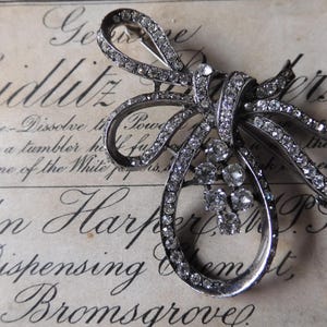Op de afbeelding: Een zilverkleurige broche in de vorm van een strik met strass. De strik is gemaakt van meerdere lussen van metaal met strass die in het metaal zijn gezet. De broche ligt op een witte achtergrond met zwarte tekst.