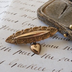 Puede incluir: Un broche vintage dorado con forma ovalada y detalles ornamentados. Un colgante en forma de corazón cuelga del broche. El broche se muestra sobre papel con escritura manuscrita, junto a un joyero de terciopelo.