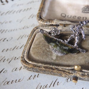 May include: A silver brooch featuring a stylized figure standing on a green stone, with a shamrock design. The brooch is in a velvet-lined box with the inscription "Queen Anne".