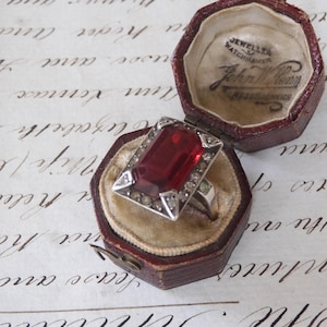 Peut inclure: Une bague vintage en argent avec une grande pierre rouge rectangulaire, entourée de petites pierres claires. La bague est présentée dans un écrin à bijoux bordeaux antique ouvert avec un intérieur beige. Le texte "JEWELLER WATCHMAKER John William" est imprimé à l'intérieur.